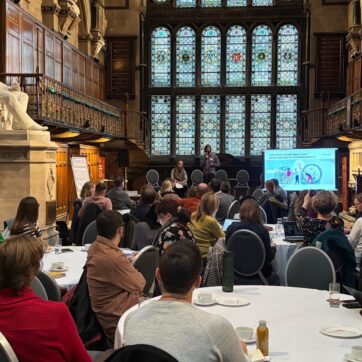 The image shows a gothic style hall with workshop participants sitting at breakout group tables following a presentation on a big screen.