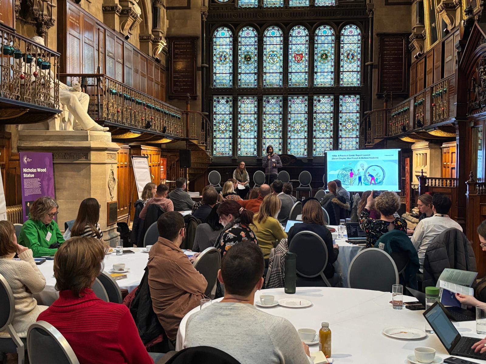 The image shows a gothic style hall with workshop participants sitting at breakout group tables following a presentation on a big screen.
