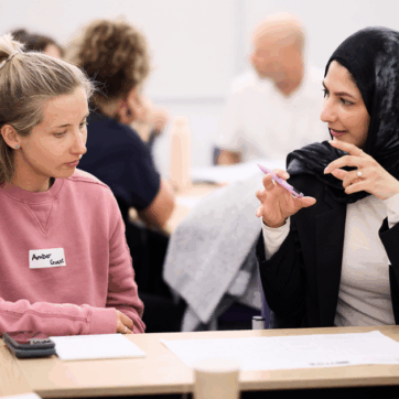 Two people are talking at a desk at the UPEN conference. One of them is wearing a black patterned headscarf and is using their hands to explain a point to the other person.