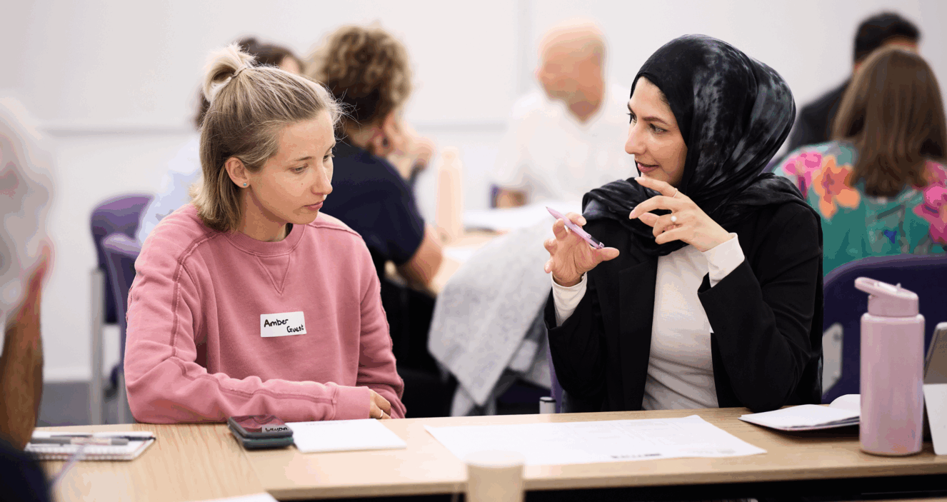 Two people are talking at a desk at the UPEN conference. One of them is wearing a black patterned headscarf and is using their hands to explain a point to the other person.