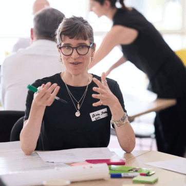 Olivia Stevenson, author of the blog, is speaking at a workshop as part of the UPEN Conference 2025. She is sitting at a table gesticulating to other workshop participants explaining something to them with post it notes in front of her.
