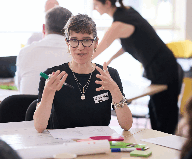Olivia Stevenson, author of the blog, is speaking at a workshop as part of the UPEN Conference 2025. She is sitting at a table gesticulating to other workshop participants explaining something to them with post it notes in front of her.