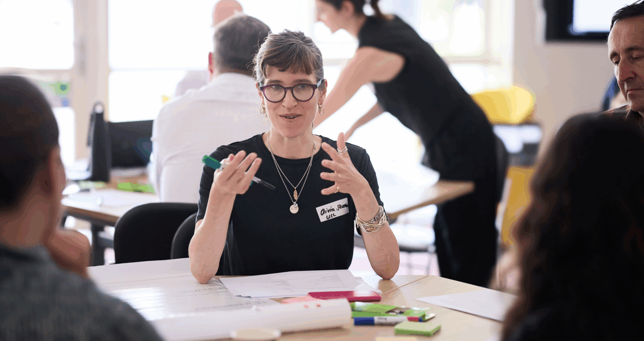Olivia Stevenson, author of the blog, is speaking at a workshop as part of the UPEN Conference 2025. She is sitting at a table gesticulating to other workshop participants explaining something to them with post it notes in front of her.