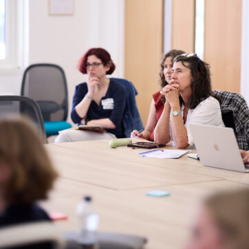 This image shows participants sitting at a large oak conference table watching a presentation (which is not in view).
