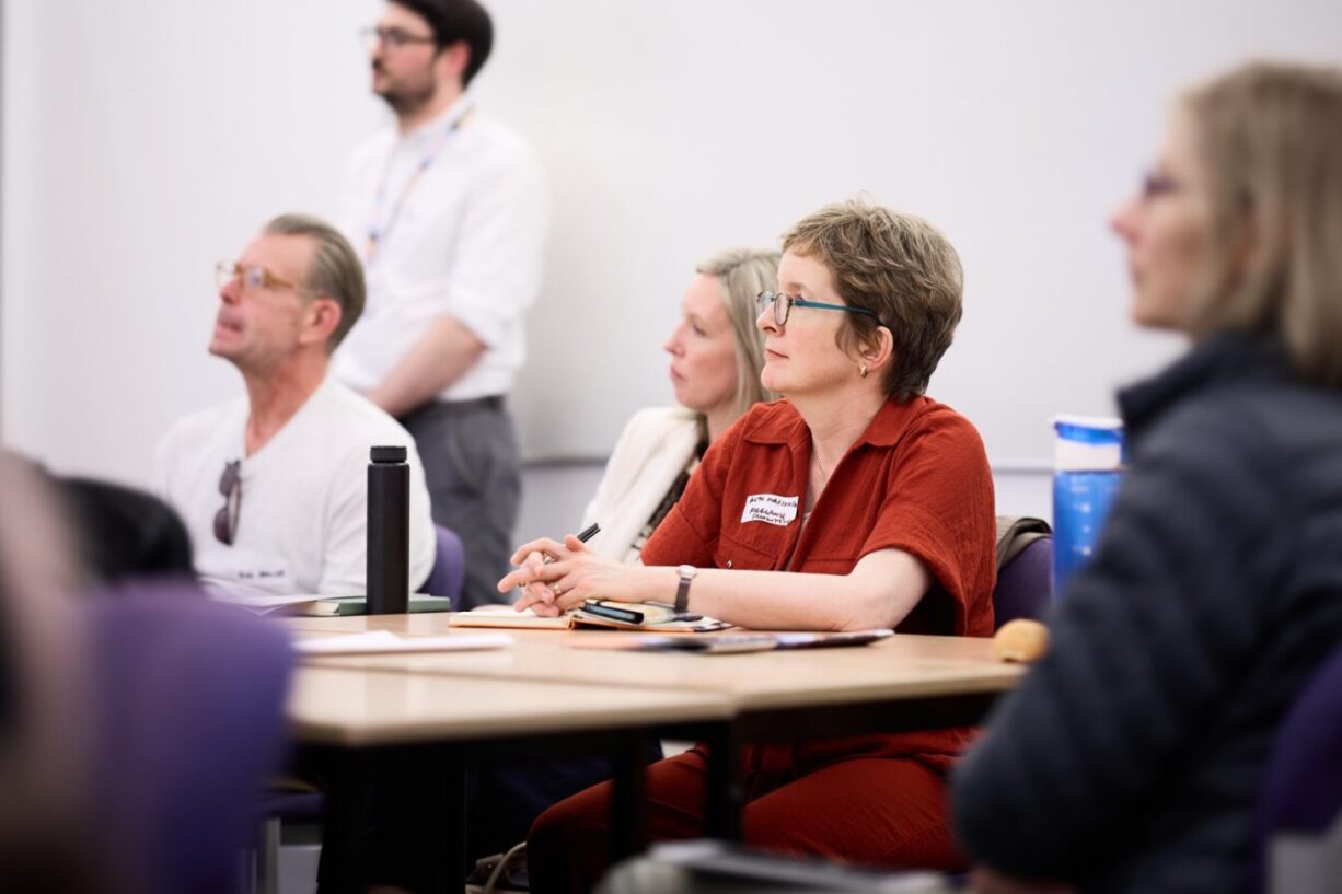 this image shows people sitting at a table looking left towards a presenter which is outside of the image. There is a white wall in the background, with a person standing next to the wall.