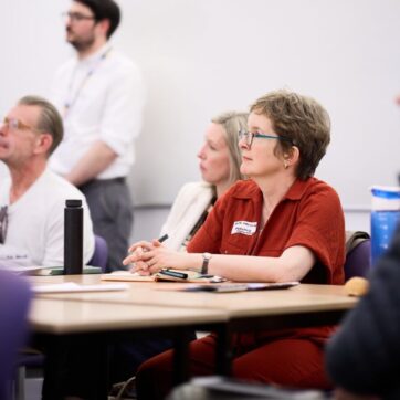 this image shows people sitting at a table looking left towards a presenter which is outside of the image. There is a white wall in the background, with a person standing next to the wall.