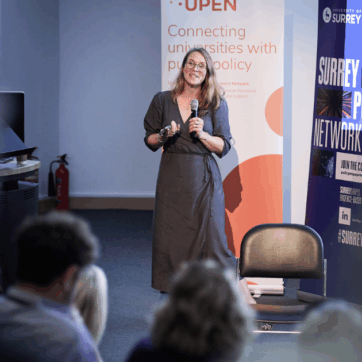 Sarah Chaytor is standing up at a conference delivering a presentation, talking into a microphone. She wears a blue dress and is standing in front of two event banners, one for UPEN and one for the University of Surrey.