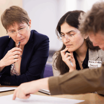 Three people, two women and a man, are sat at a table looking at a document they appear to be jointly working on together. The man is pointing at something on the page while the women are looking in concentration at the document.