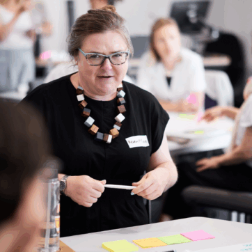 The image shows people gathered at a workshop, with three people in the foreground sat at a table. A woman is explaining something to the table and holding a pen in her hand. The table has post its and documents on it.