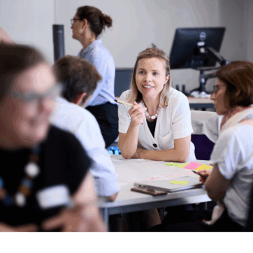 Background image of a classroom with a computer monitor in the background as well as a person facing the back. In the foreground are blurry sillhouettes of two people engaged in conversation. The focus of the image is on a woman with blonde hair engaged in conversation and smiling.