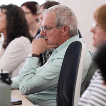 This image shows people sitting at a table looking towards a screen that is not in view. The background is white with a window.