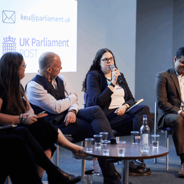 This image is of the thematic research leads session at the UPEN 2025 conference. Four people are in shot sitting on chairs in front of a glass table with glasses of water on it. They sit in front of a big screen with the logo of the UK Parliament on it. One person is holding a microphone and speaking into it while the others listen.