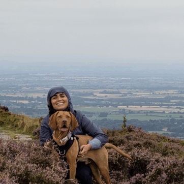 A photo of Azadeh Fatehrad who is kneeling at the top of a hill covered in purple heather with fields in the background. She is smiling and hugging a caramel coloured dog who is standing and looking into the distance.