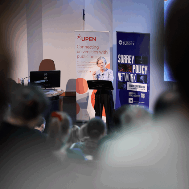 Professor Dame Angela McLean gives a conference presentation standing in front of a lecturn with banners for UPEN and the University of Surrey behind her. There are people out of focus in the foreground of the image in the audience.