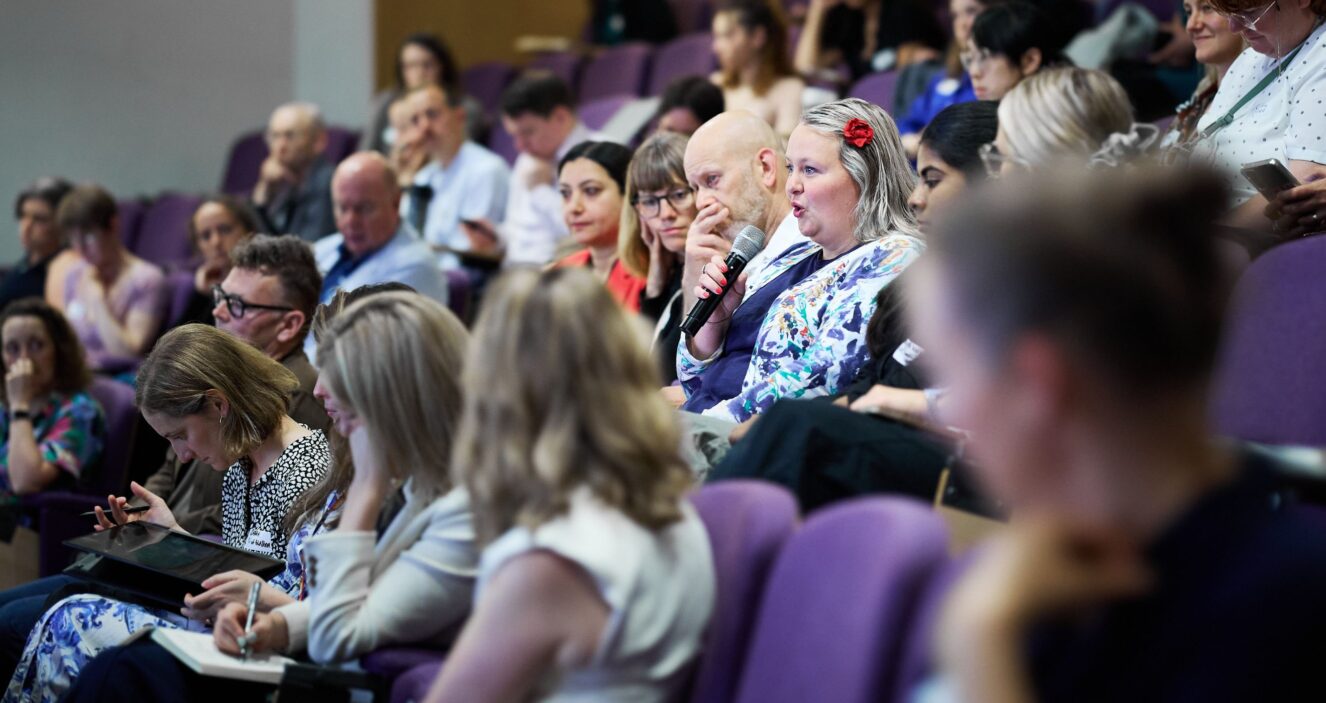 An image side on of a conference theatre showing the audience only, sitting in purple seats. A woman wearing a patterned top and a flower in her hair is holding a microphone and asking a question.