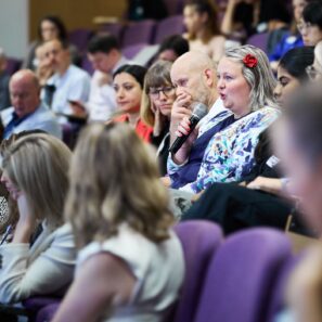 An image side on of a conference theatre showing the audience only, sitting in purple seats. A woman wearing a patterned top and a flower in her hair is holding a microphone and asking a question.