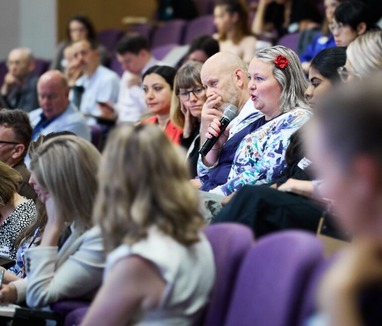 An image side on of a conference theatre showing the audience only, sitting in purple seats. A woman wearing a patterned top and a flower in her hair is holding a microphone and asking a question.