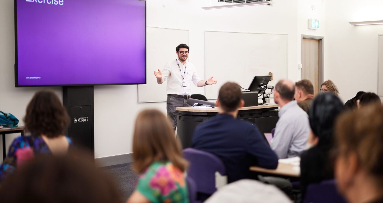 A man is standing in front of a desk giving a presentation to a room full of people in a workshop. He has open arms as he is talking to them.