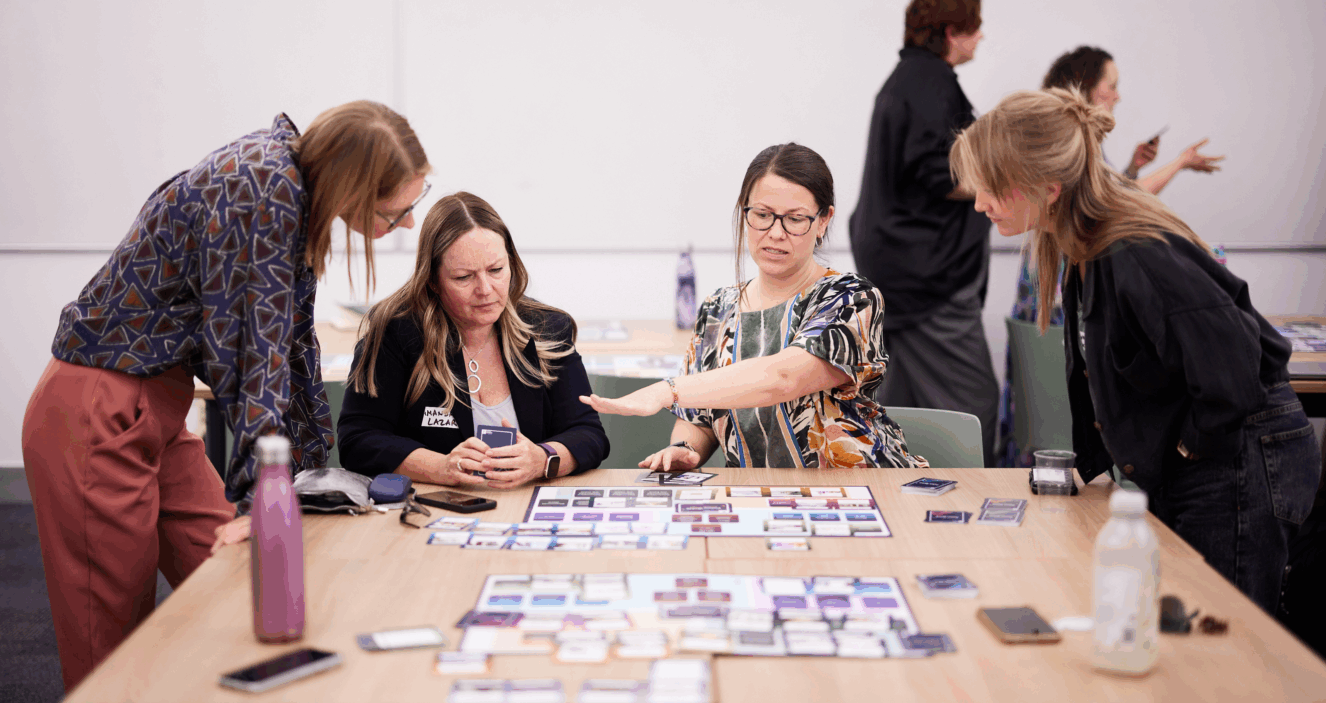 Four people are around a table, the two on the furthest left and right are leaning over, and two people are sat facing the camera. They are looking at a spread of information on the table in front of them. One of then is gesticulating with their hand and talking to the others.