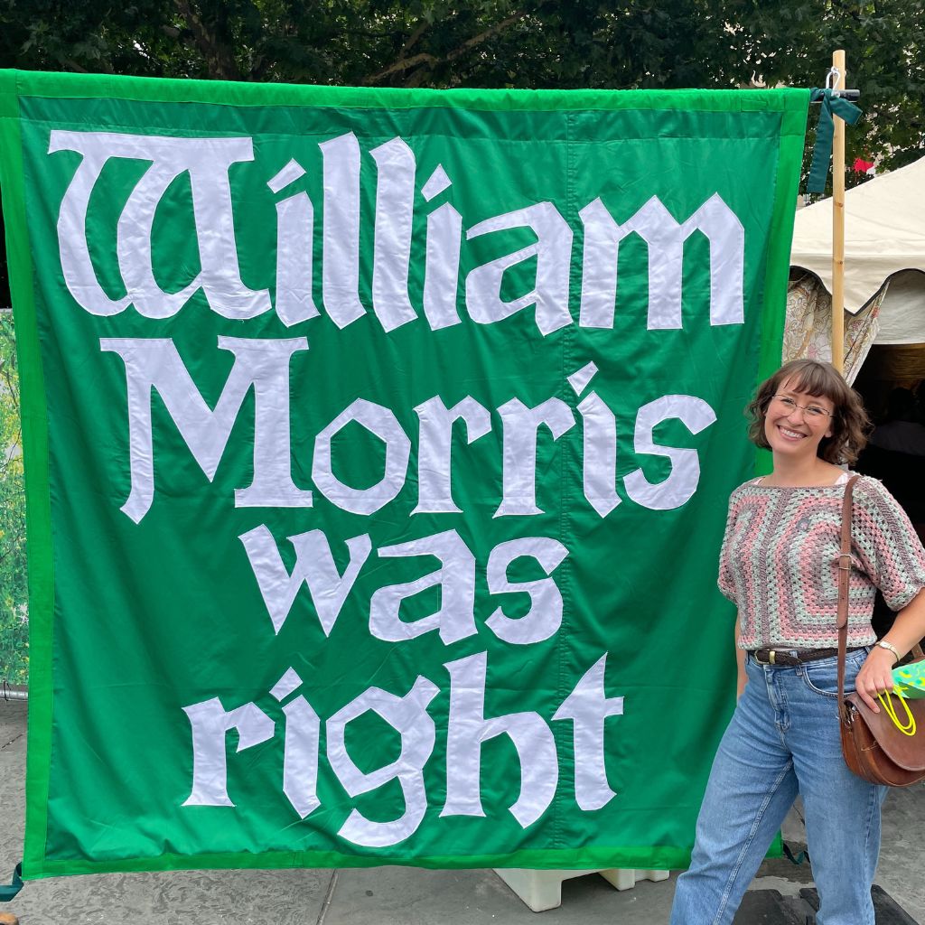 A person stands to the right of a big green banner reading "Willian Morris was right" in white. She is facing the screen and smiling, wearing blue jeans and a crochet top.