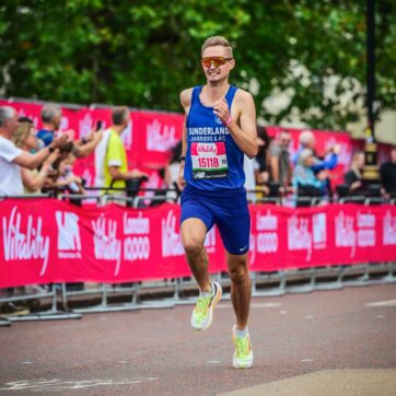 Joe Armstrong is pictured running a race, mid stride. He wears blue sports clothes and has yellow running trainers with reflective red sunglasses. Behind him is a red banner with Vitality written on it with lines of spectators.