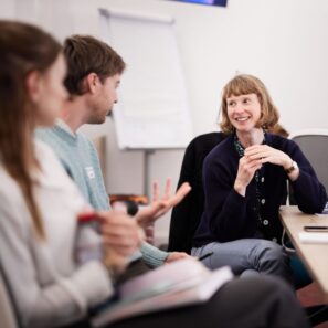 A photo of three people at a conference workshop. The camera focuses on one woman who is smiling and has her hands clasped listening to another one of the three make a point. They are seated around the side edge of a table.