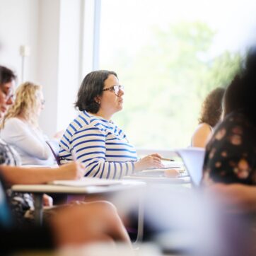 This image shows a person sat at a desk in a classroom looking towards the front of the room.
