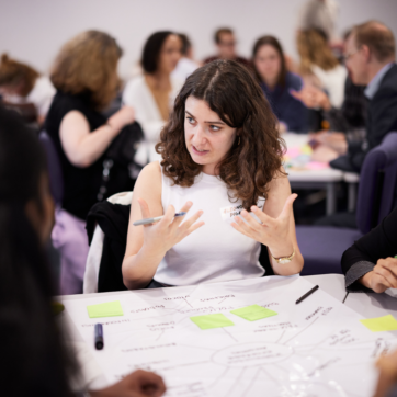 A woman sits at a table covered in post it notes at a conference table. She is explaining something to others on the table gesticulating using her hands. She wears a white sleeveless top, and you can see other conference participants out of focus in the background.