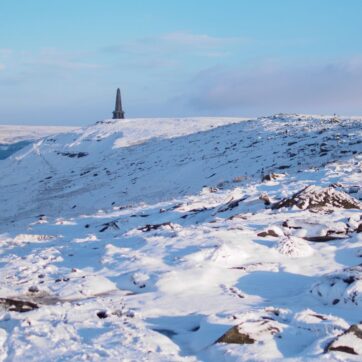 Snowy hills with a light blue sky and scant clouds. In the distance an obselisk stands on the horizon.