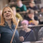 A photo of Mona Krewel giving a lecture. She is in focus in a blue shirt gesturing to a screen with the audience out of focus behind her.