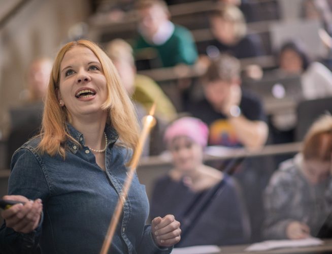 A photo of Mona Krewel giving a lecture. She is in focus in a blue shirt gesturing to a screen with the audience out of focus behind her.