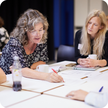 People working together round a desk, with a woman leaning forward to write on paper.