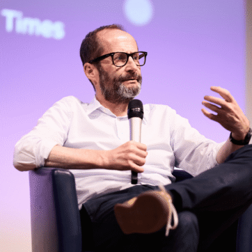 This image shows Alan Smith delivering the keynote at the UPEN Conference. They are sitting on a chair on the stage holding a microphone and talking to the audience.