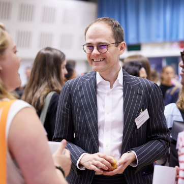 A person in a suit is standing up, and smiling off camera to someone they are talking to. Other people can be seen in the background networking.