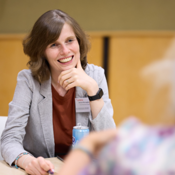 Image shows someone in a light grey blazer with a red top and brown shoulder length wavy hair sat at a workshop table. They are smiling and looking at the person opposite them who is slightly off camera in the foreground to the right.