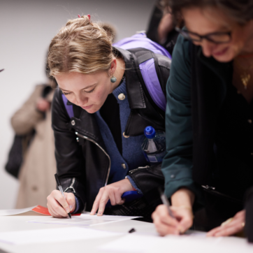 A person is leaning forward to write on a desk with their right hand. They have light brown hair tied up and a purple backpack. There is someone on the right of the frame doing the same, but out of focus. They appear as if they are reigstering for a workshop or event.